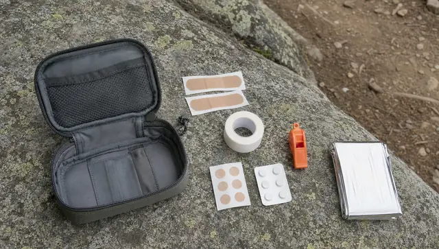 A small open grey first aid pouch on a lichen-covered rock with bandages, tape, blister patches, an orange whistle and a foil blanket