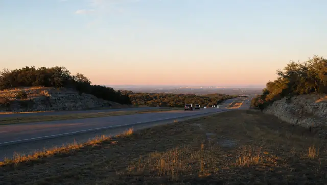 Early dusk on a divided highway running east through Texas Hill Country, live oaks along the verge and a few cars heading toward the glow of a city