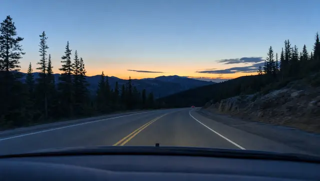 A two-lane mountain highway at dusk seen through a car windshield, with the last orange light over the ridge
