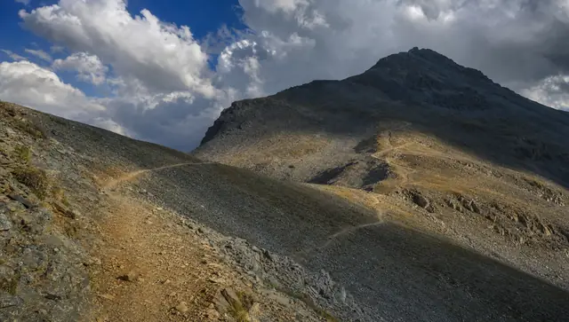 A rocky alpine trail switchbacking up a bare slope toward a summit as afternoon clouds build overhead