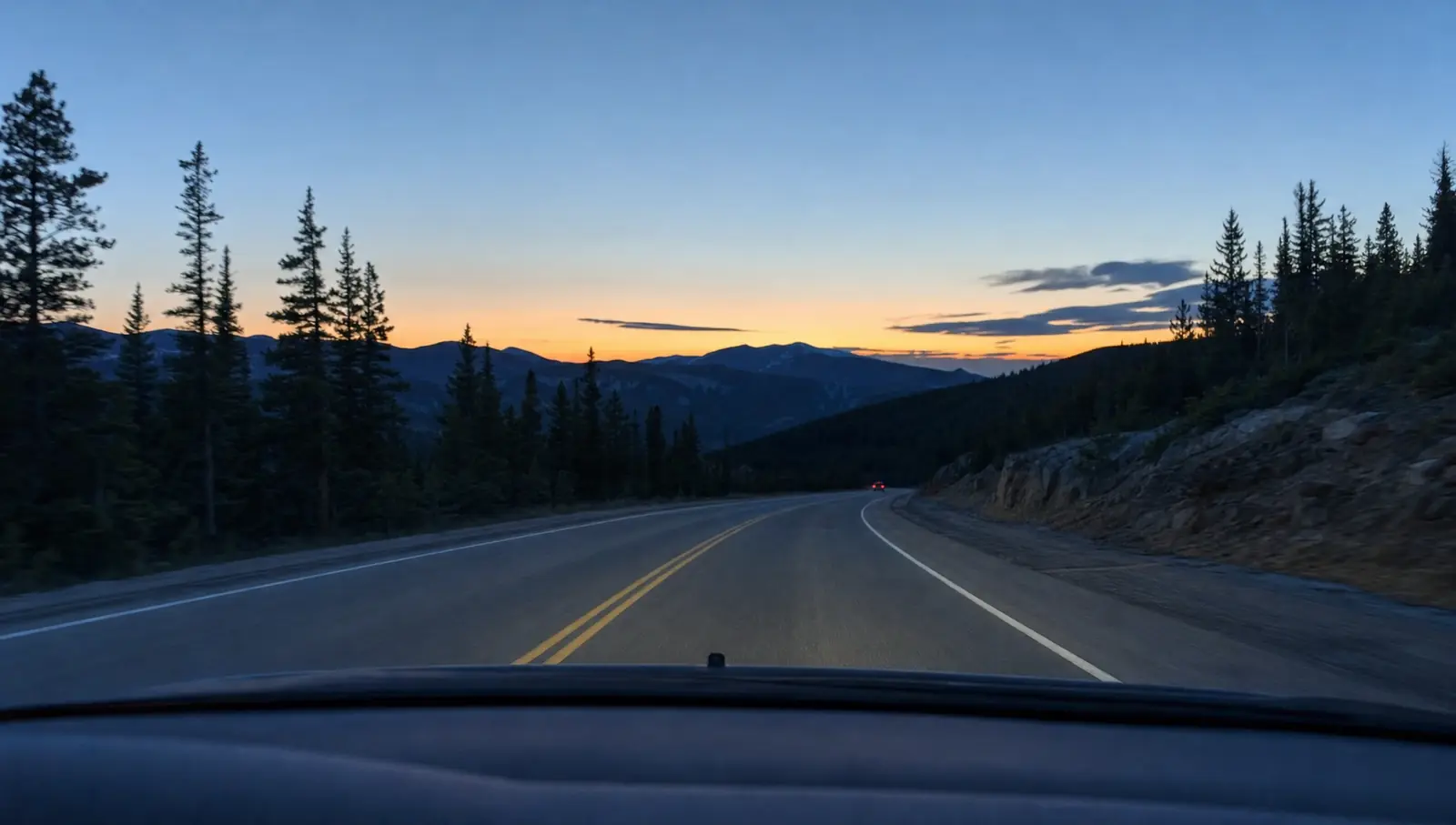 A two-lane mountain highway at dusk seen through a car windshield, with the last orange light over the ridge